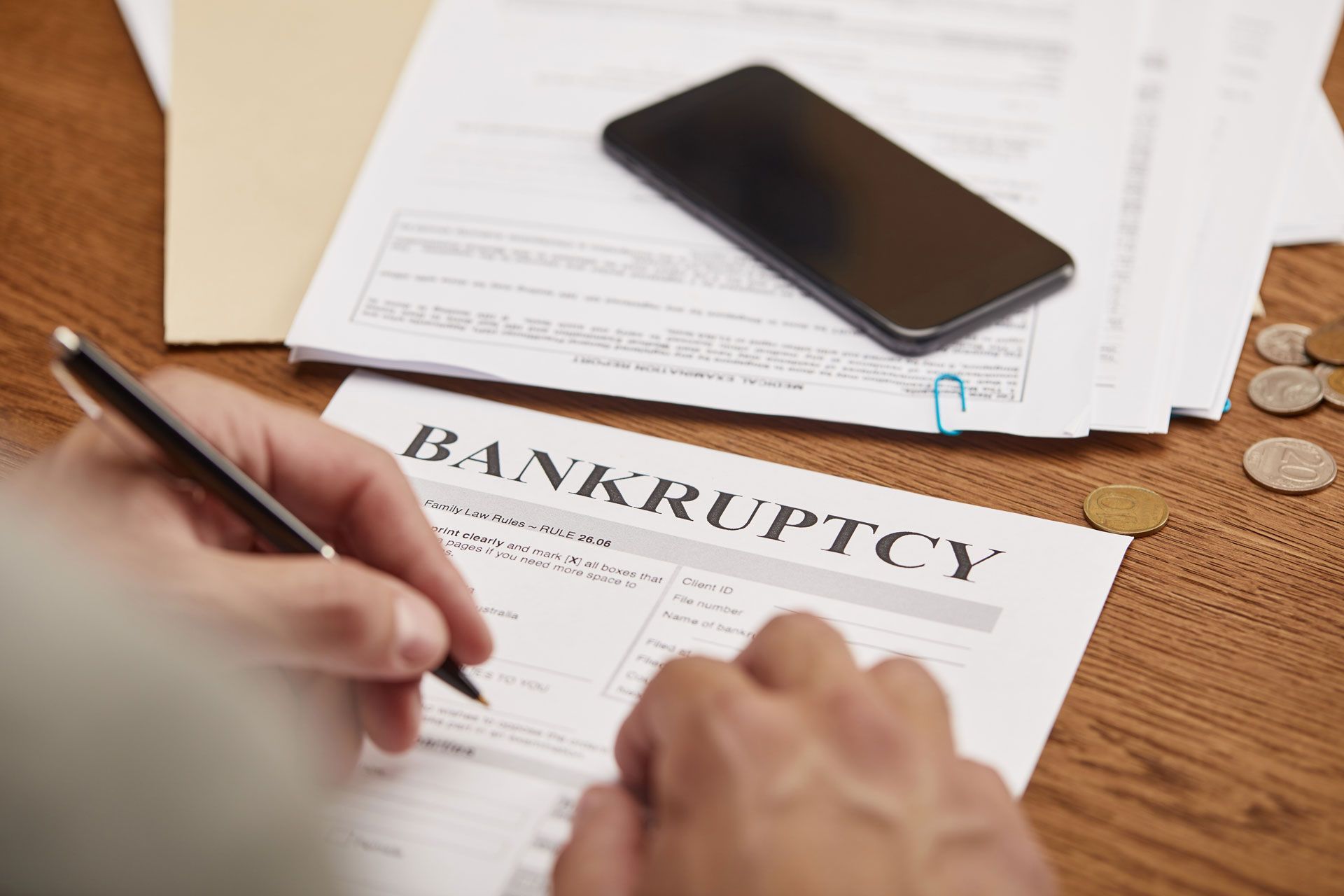 Hands signing bankruptcy form beside a smartphone on a desk with documents and coins