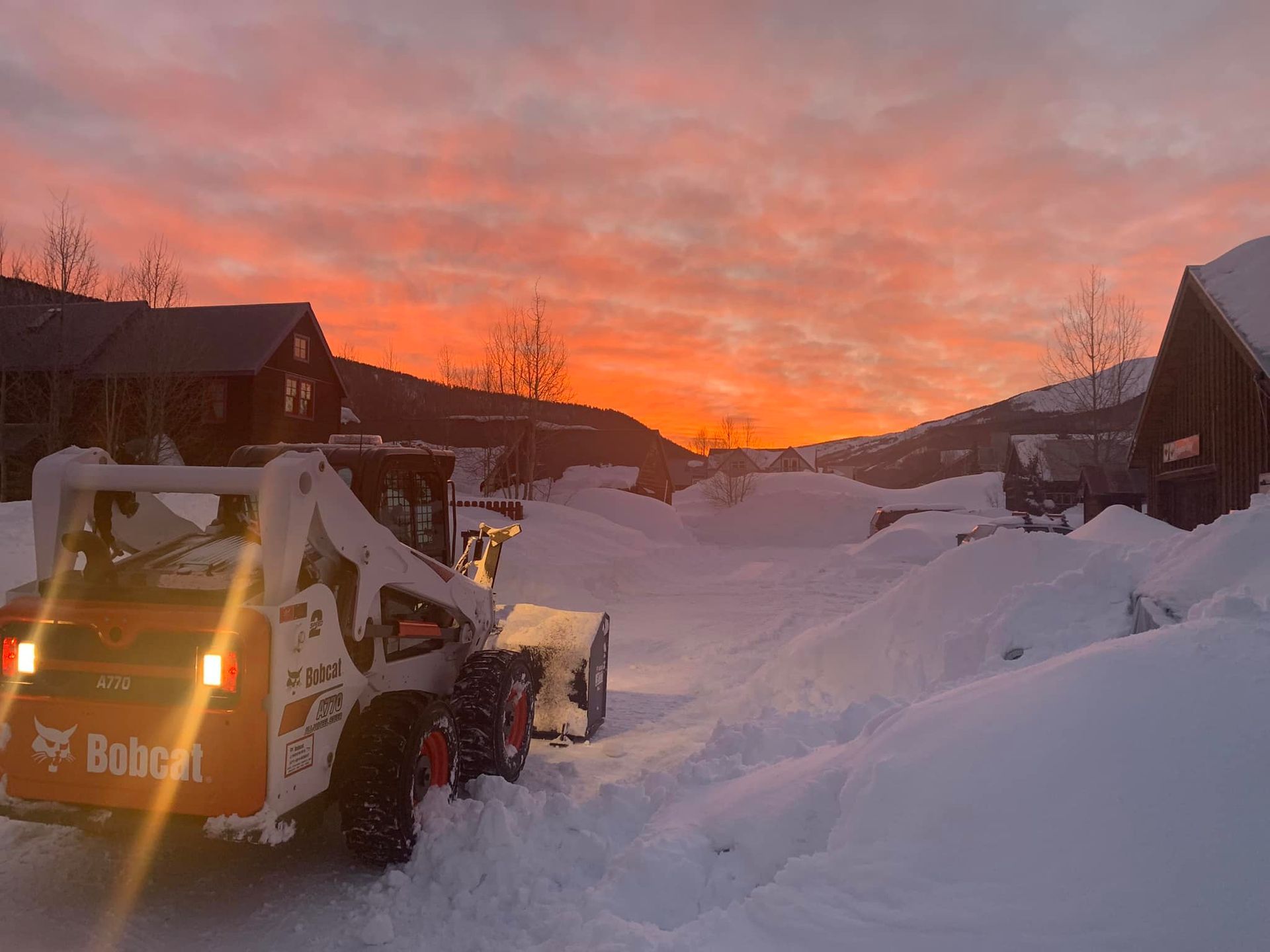 A snow plow is driving down a snow covered road at sunset.