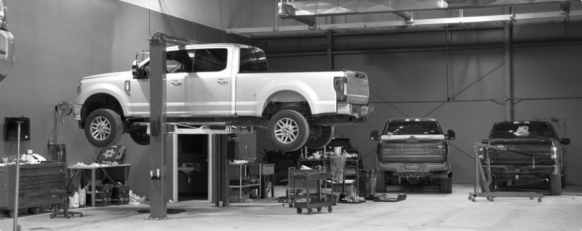 White SUV on a lift in an auto repair shop with several vehicles parked below | G3 Transmission and Auto
