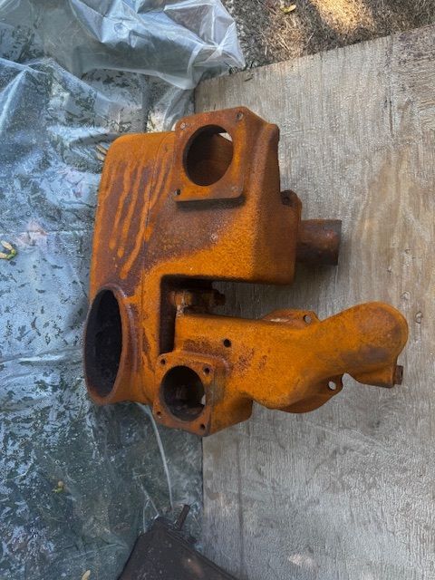 Rusty, orange-brown metal machine housing with various holes and a cylindrical projection, lying on wood and cloth.