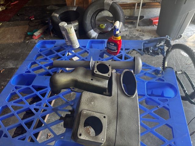 Gray painted machine parts on a blue plastic pallet, with spray cans, tires, and a bicycle wheel in a garage.