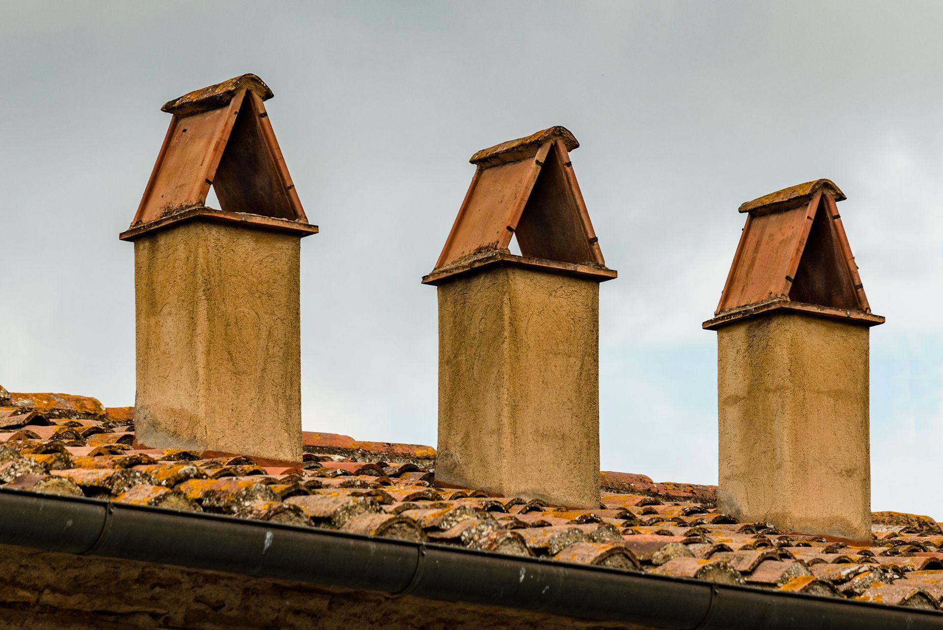 Three weathered rooftop chimneys with terracotta tops on a tiled roof against a cloudy sky