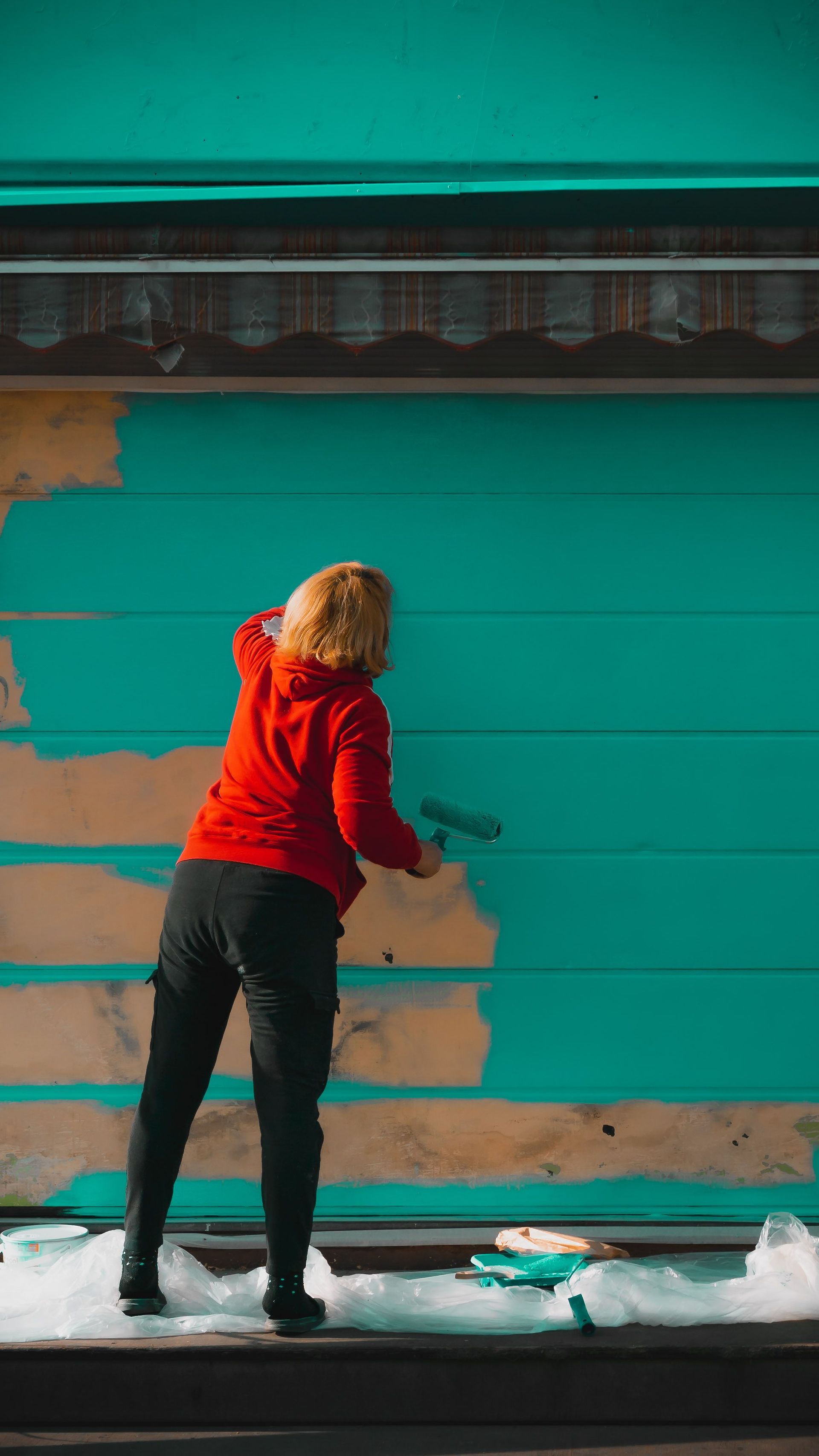 Person painting a turquoise wall with a roller near peeling paint, standing on a snowy ledge.