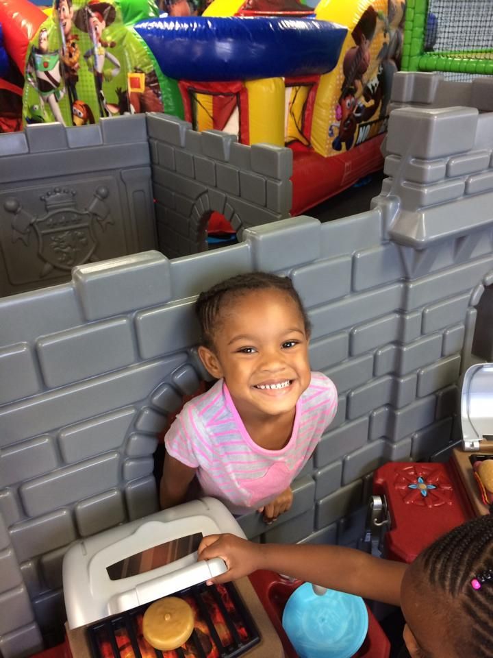 A little girl is playing in a playhouse with a bouncy house in the background.