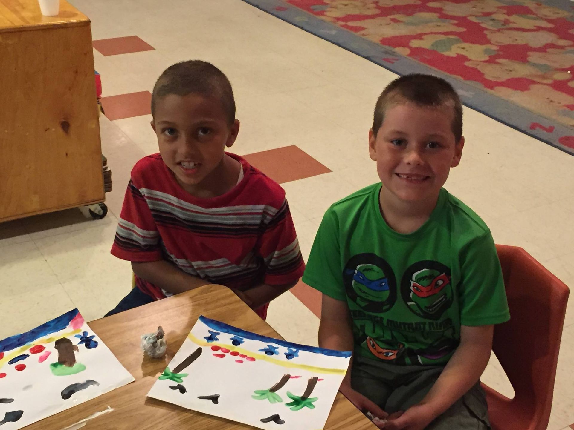 Two young boys are sitting at a table with paintings on it