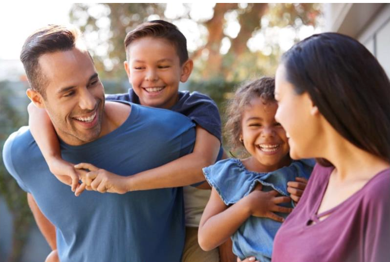 A man is carrying two children on his back while a woman looks on.