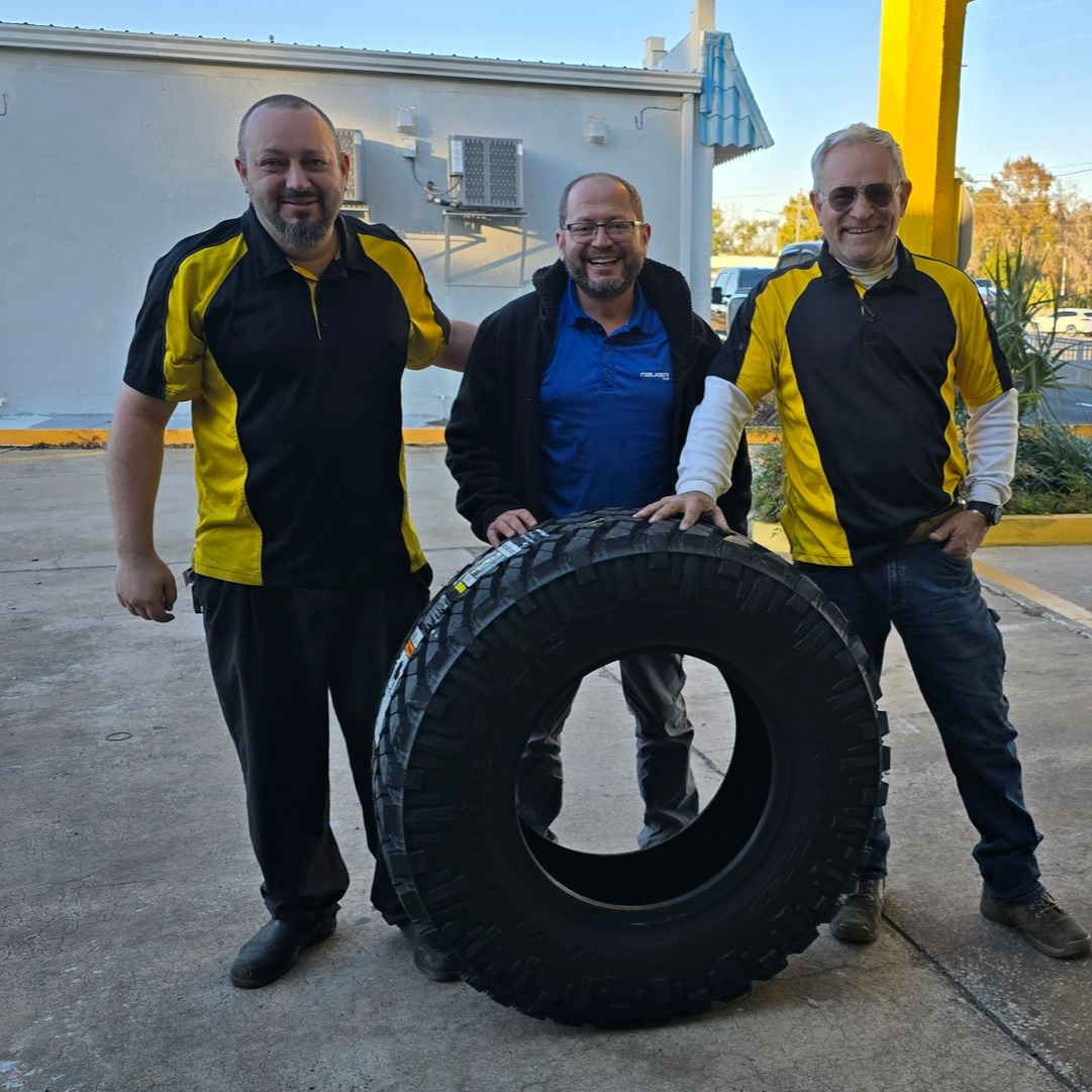 Three men pose with a large tire outdoors. Two wear yellow/black shirts. One wears a blue shirt and jacket.