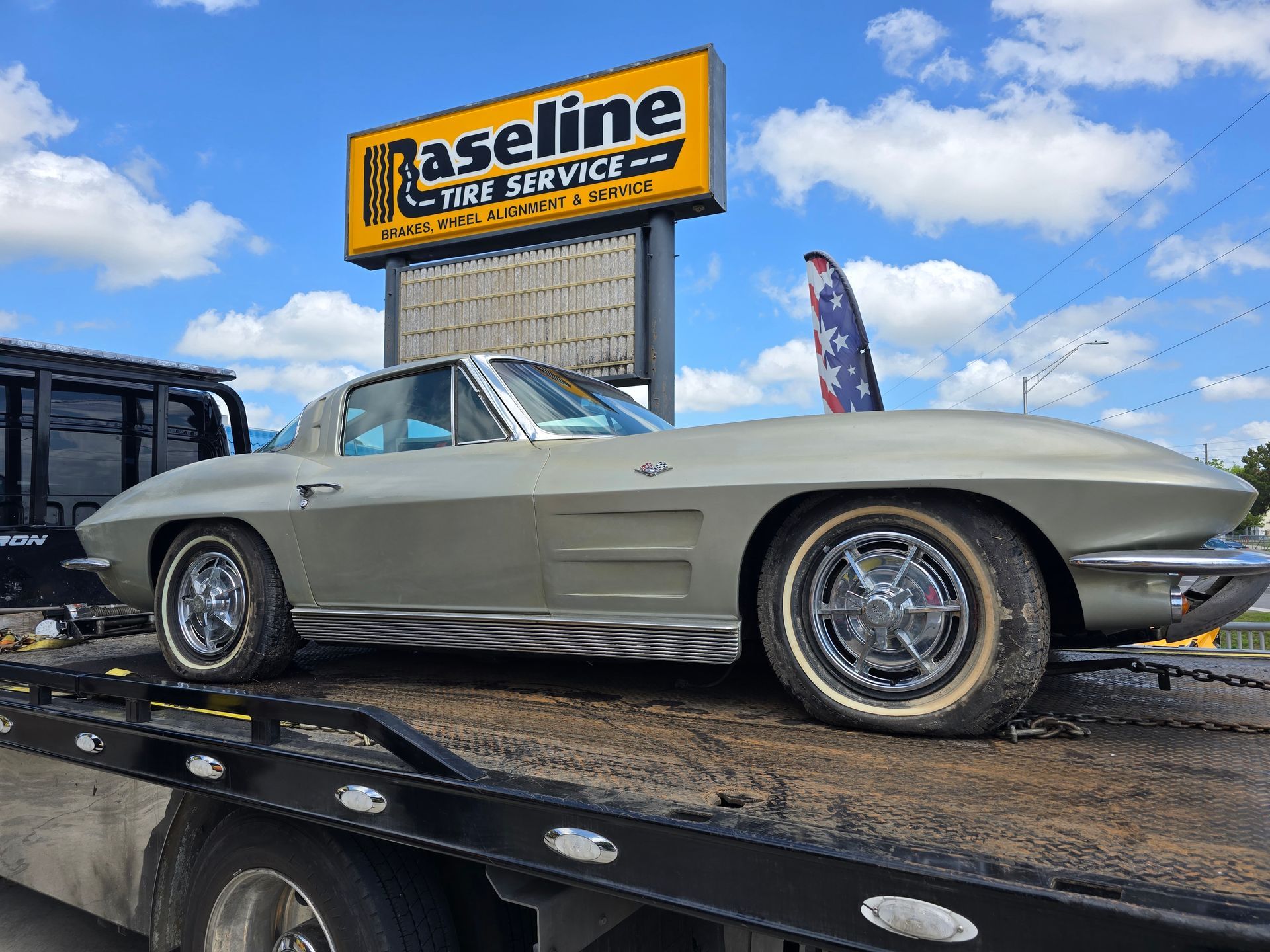 Silver vintage Corvette being towed by a flatbed tow truck in front of 