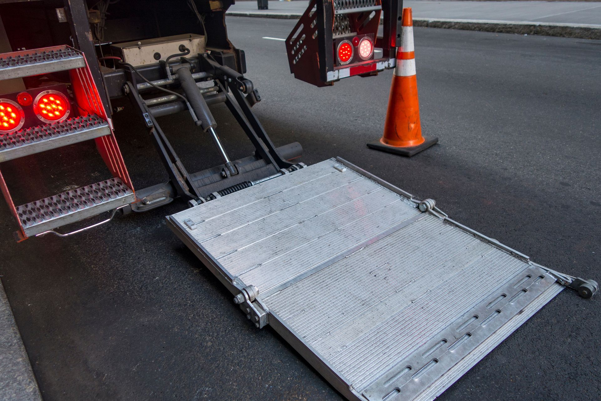 A truck's lowered hydraulic lift. Orange cone on the street.