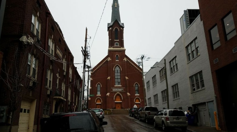 Red-brick church with tall steeple on a narrow city street, flanked by parked cars and buildings