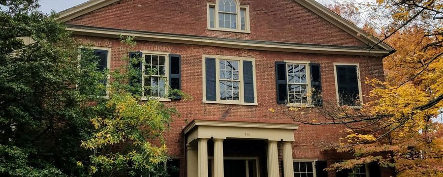 Red brick house with white columns and shutters, partly framed by trees in autumn colors