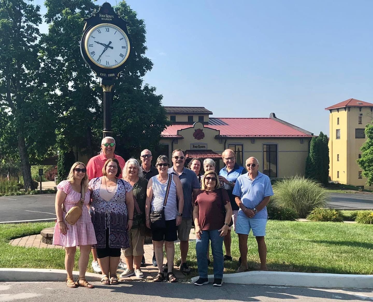 Group of people posing outdoors by a large clock in front of buildings on a sunny day