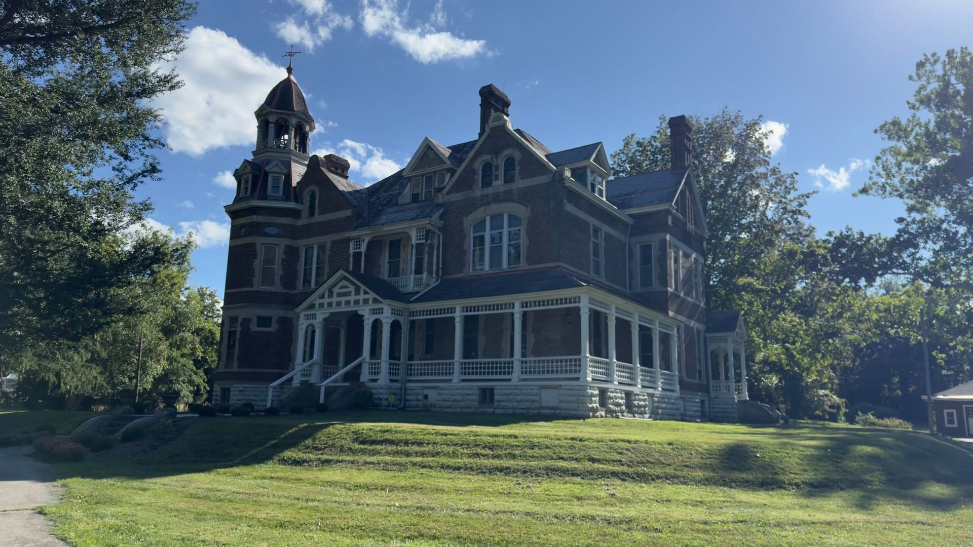 Victorian-style mansion with dark trim and white porch, set on a grassy hill under a blue sky with trees.