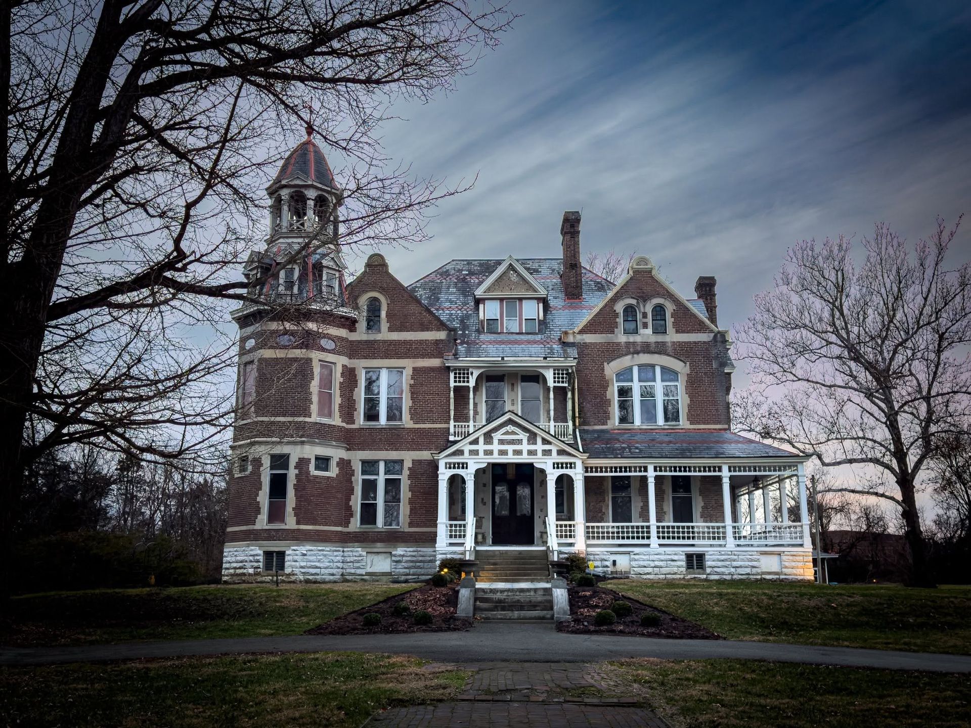 Large Victorian-style brick house with a tower, white porch, and bare trees under a cloudy sky