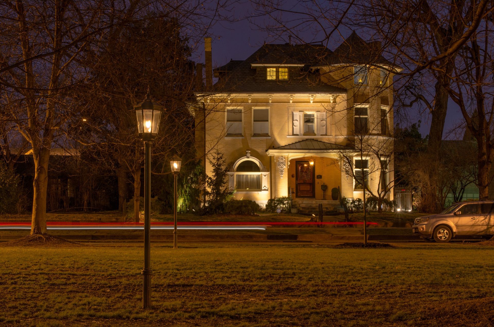 Lit house at night with warm windows, trees, a streetlamp, and parked cars in front.