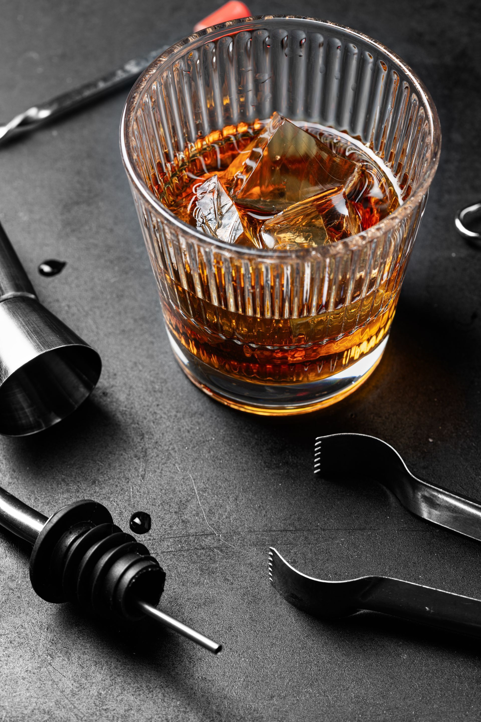 Glass of whiskey with ice on a dark table, surrounded by cocktail tools and a black pepper mill.