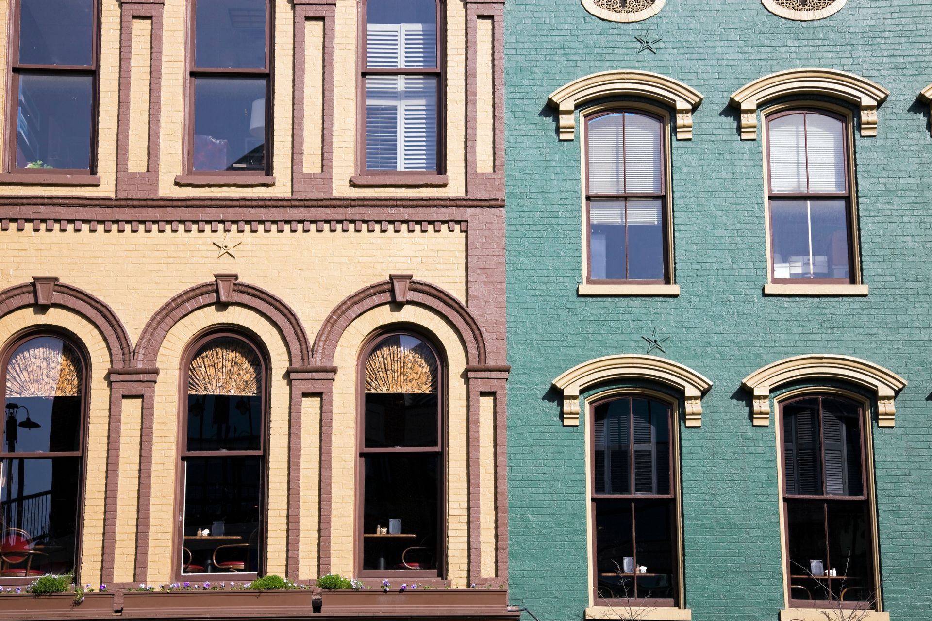 Two adjacent historic building facades, one tan brick and one green-painted, with arched and bay windows.
