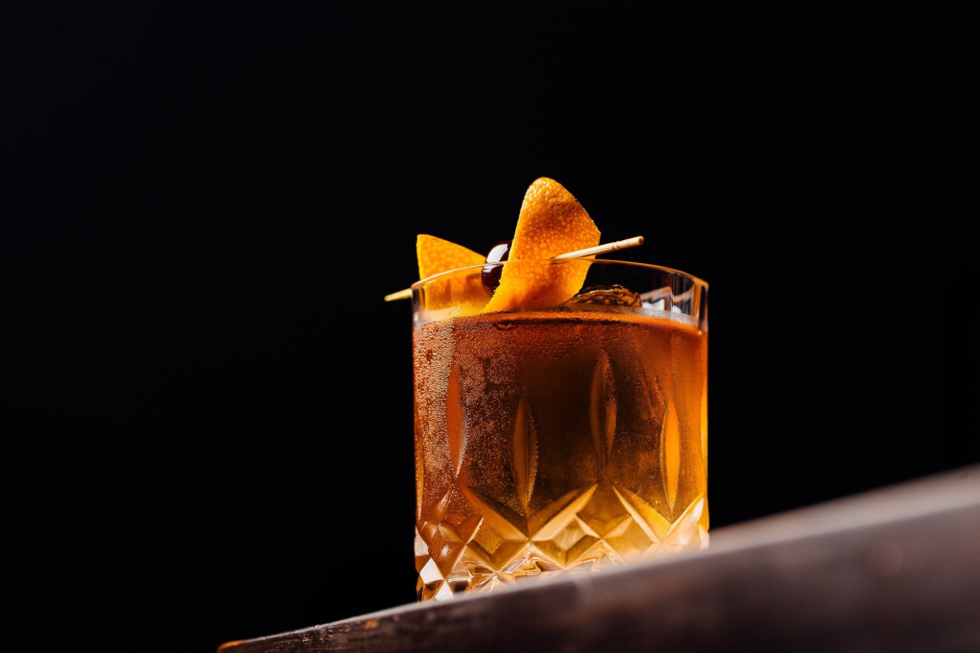 Amber cocktail in a faceted glass with orange garnish on a dark bar top