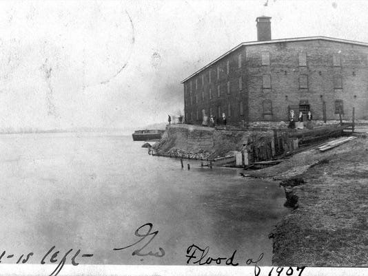 Historic black-and-white waterfront scene with a large brick building on a shoreline dock.