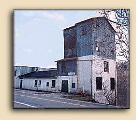 Old white industrial building with a tall weathered tower beside a road