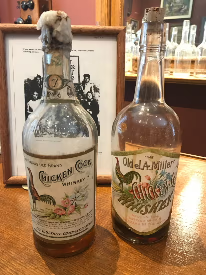 Two vintage liquor bottles on a wooden table, one with a cork and one labeled Old L. Miller Whiskey.