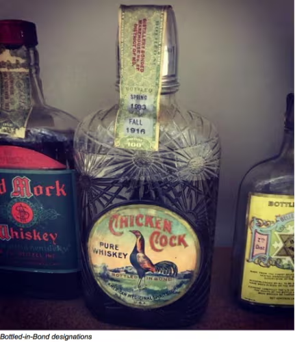 Three vintage whiskey bottles on a shelf, including a central “Chicken Cock” bottle and a “Red Mark Whiskey” bottle.