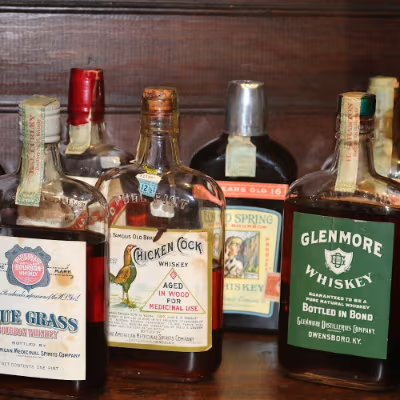 Assorted vintage liquor bottles on a wooden shelf, including Glenmore whiskey and Chicken Cock bourbon labels