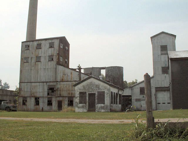 Abandoned industrial buildings with tall smokestacks on a grassy lot under a cloudy sky