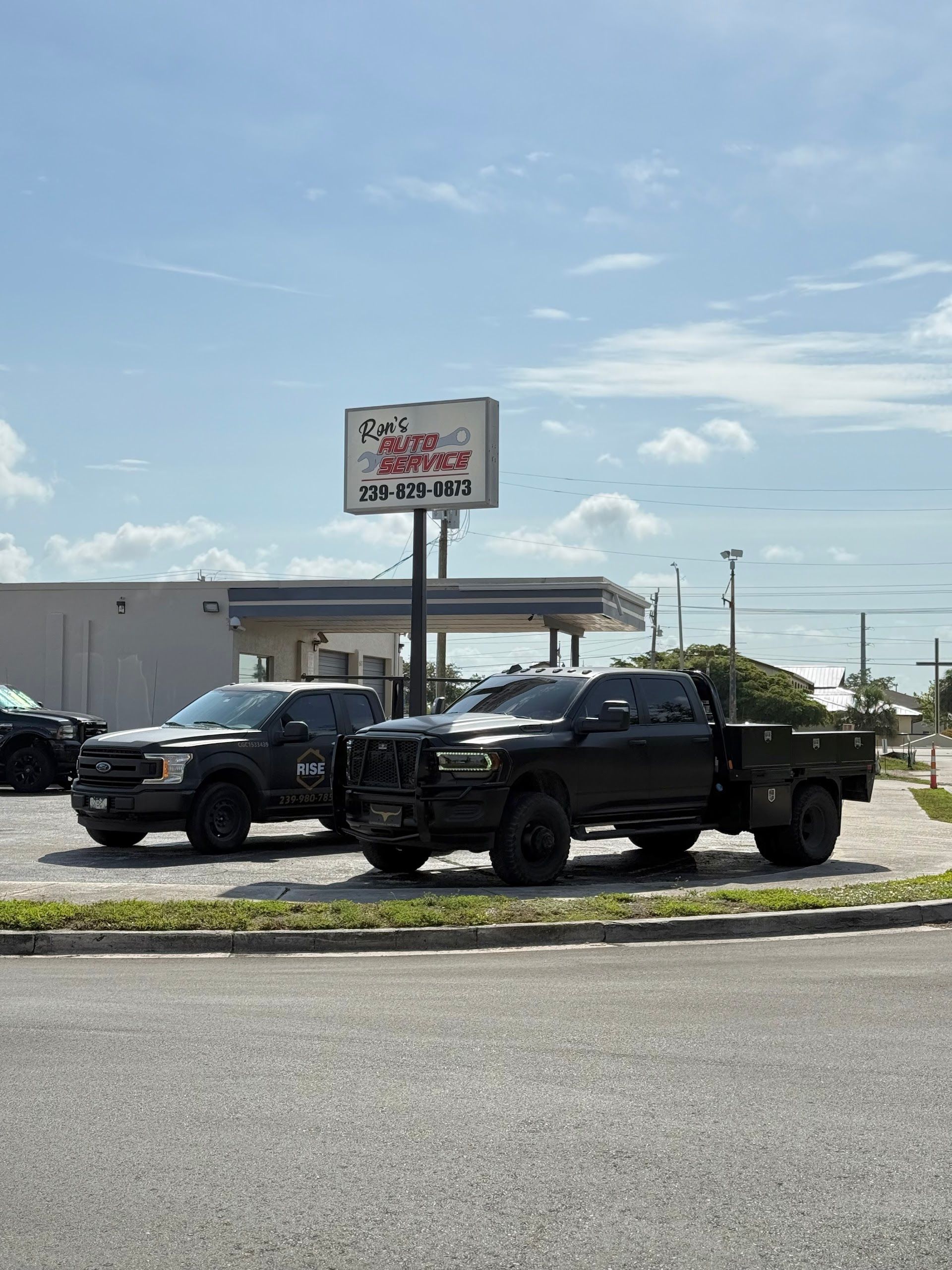Two black pickup trucks parked in front of a roadside sign under a partly cloudy sky. | Ron's Auto Service