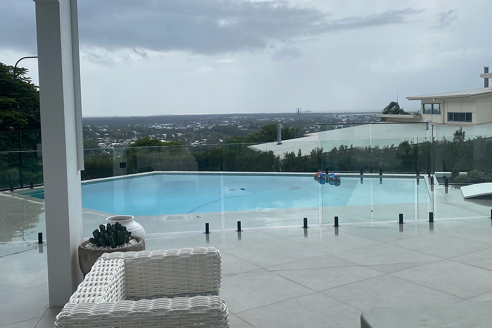 A Pool with a Glass Fence Overlooks a City Under a Cloudy Sky — Frameless Shower Installations In Maroochydore, QLD