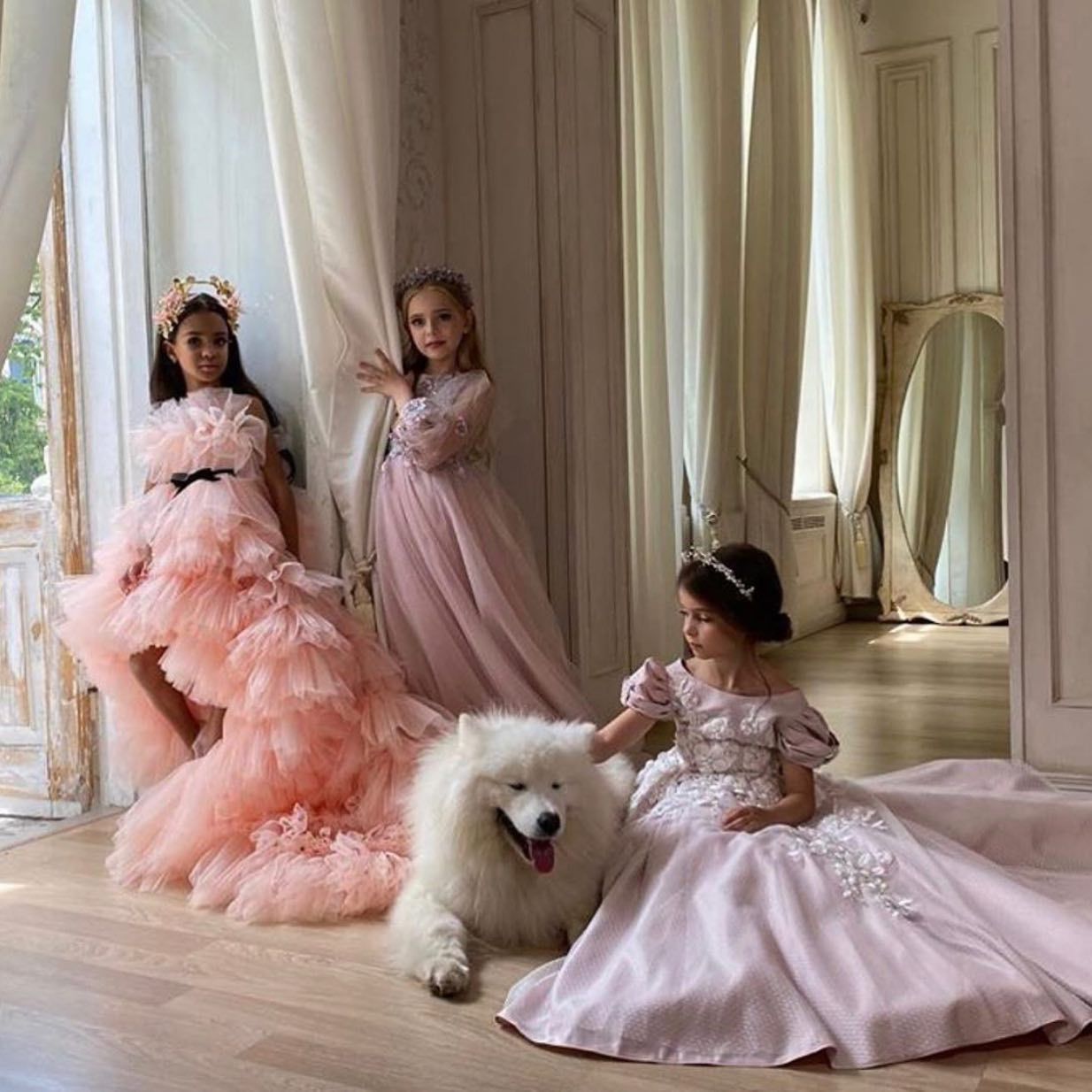 Three little girls in pink dresses are sitting next to a white dog.