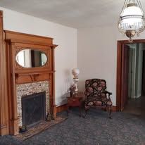 Vintage room with ornate wooden fireplace, patterned chair, carpet, and doorway to a dim hall.