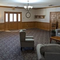 Empty carpeted room with wooden paneling, chairs, table, wall clock, and chandelier