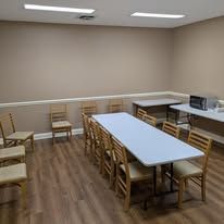Empty beige break room with wooden chairs, long white tables, and a kitchenette counter