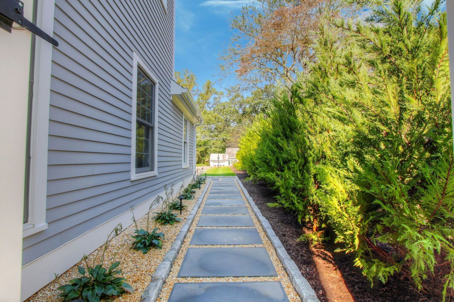 Side yard walkway with stepping stones beside a gray house and tall green hedges
