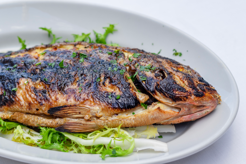 Pesce intero grigliato su un piatto bianco con guarnizione verde.