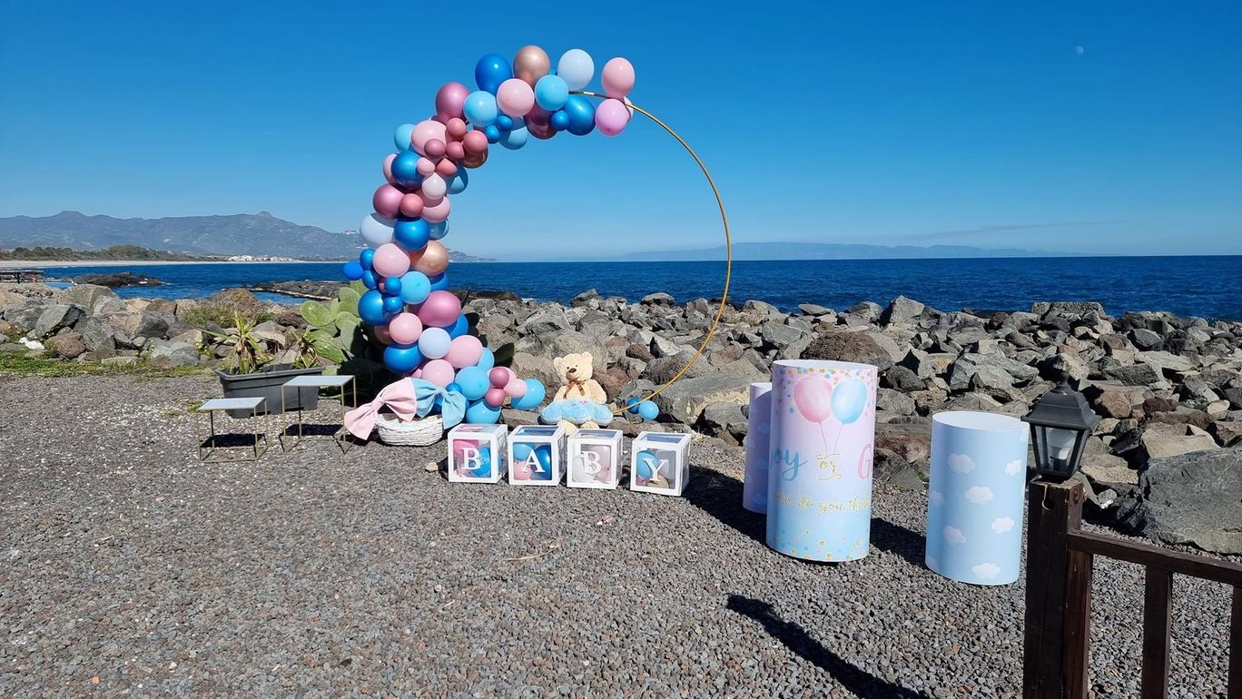 Allestimento per baby shower in riva al mare con palloncini blu, rosa e oro, un arco rotondo e scatole decorative.