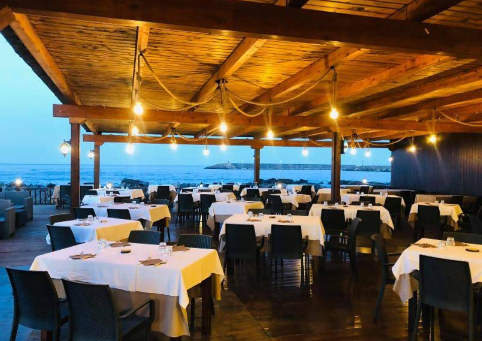 Ristorante all'aperto con tavoli apparecchiati per la cena, con vista sul mare. Luci a ghirlanda illuminano il soffitto con travi in ​​legno.