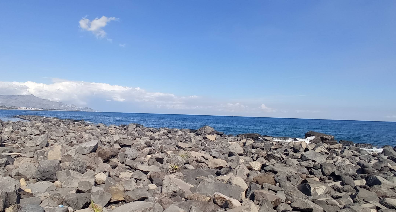 La costa rocciosa incontra l'oceano blu sotto un cielo azzurro brillante con nuvole sparse.