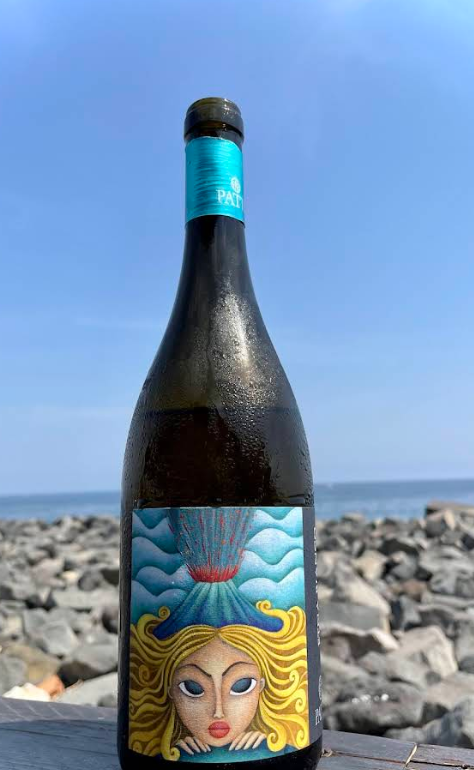 Bottiglia di vino con etichetta raffigurante un volto di donna e un vulcano blu sullo sfondo di un cielo azzurro e dell'oceano.