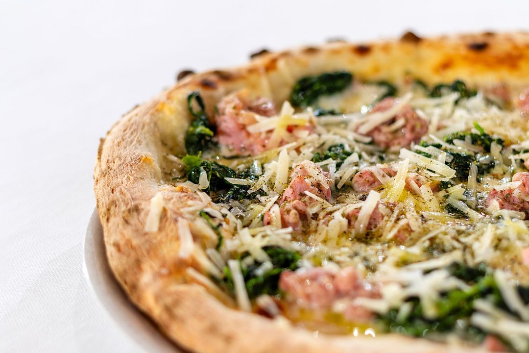 Pizza con salsiccia, spinaci e formaggio grattugiato, su un piatto bianco.