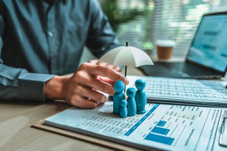 Hand placing tiny figures under a small umbrella beside a laptop and finance charts on a desk