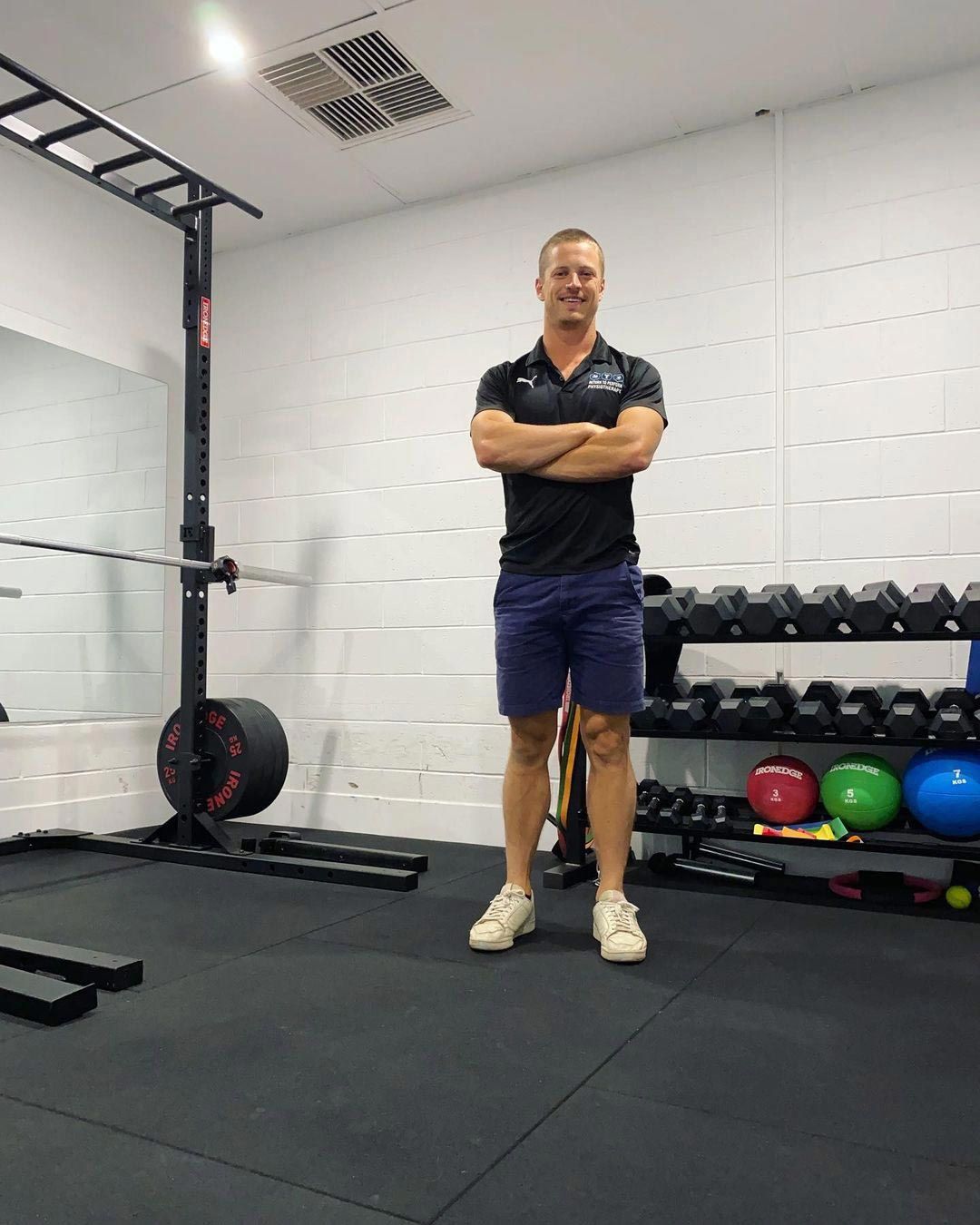 Person standing with arms crossed in a gym beside dumbbells and exercise equipment.
