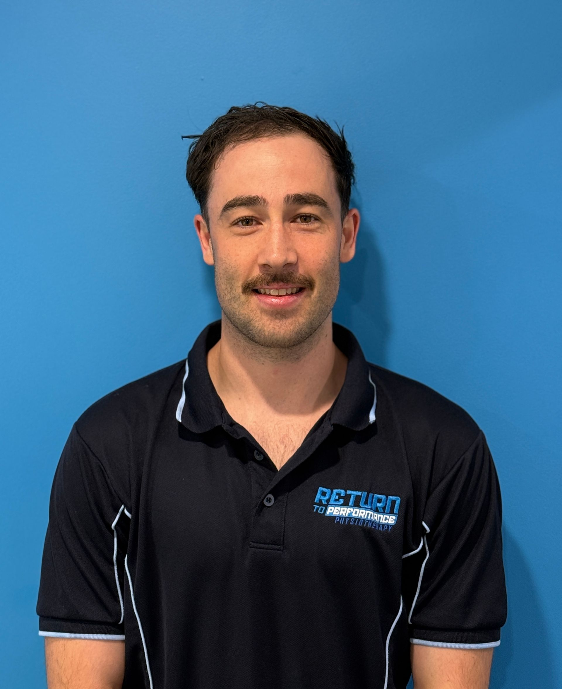 A Smiling Person With a Mustache Wearing a Black Collared Work Shirt — Return to Performance Physiotherapy in Epsom, VIC