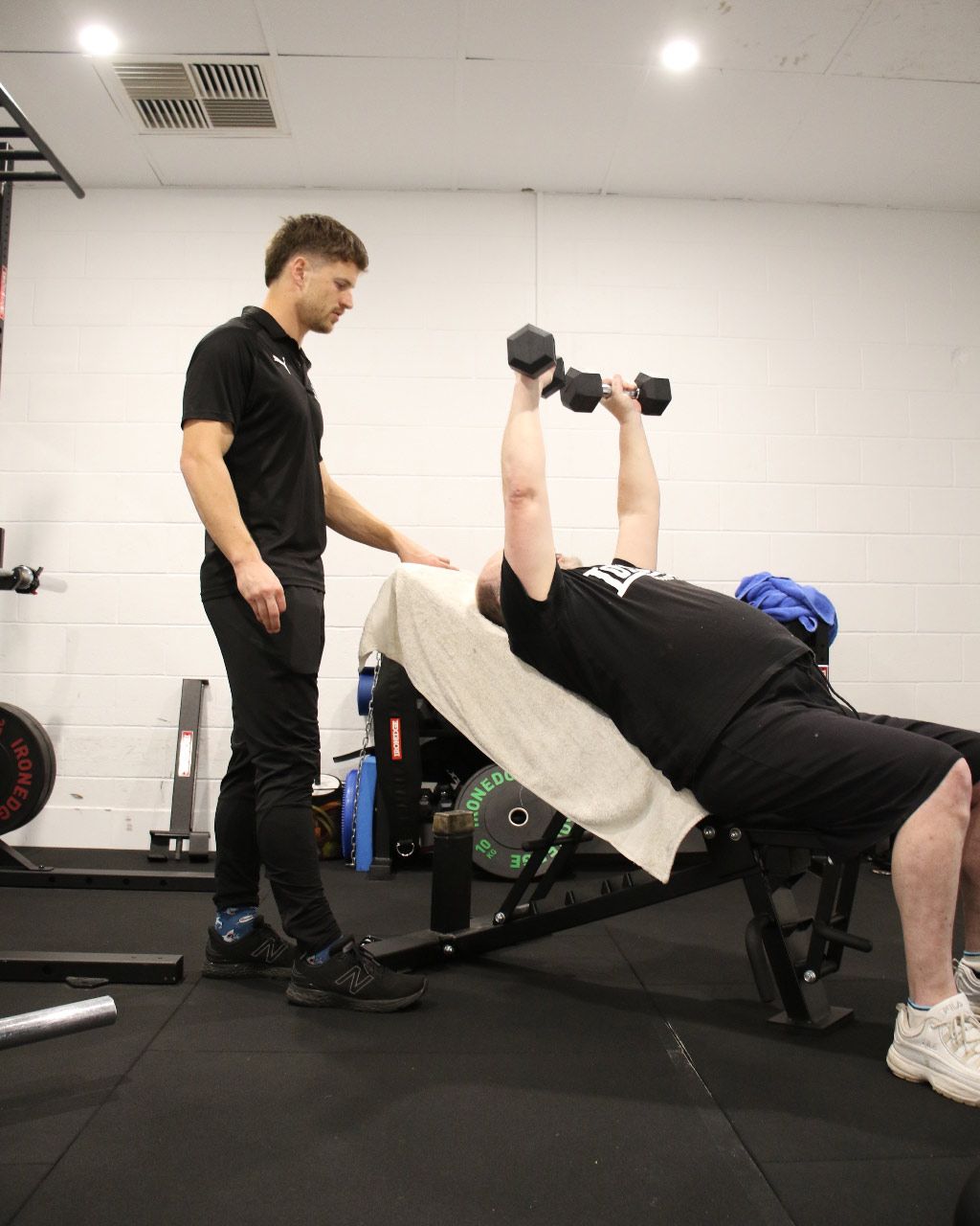 Person doing dumbbell bench press while a trainer spots in a gym