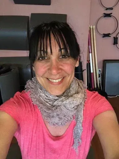 Smiling person in a pink shirt and gray scarf, seated indoors against a pink wall.