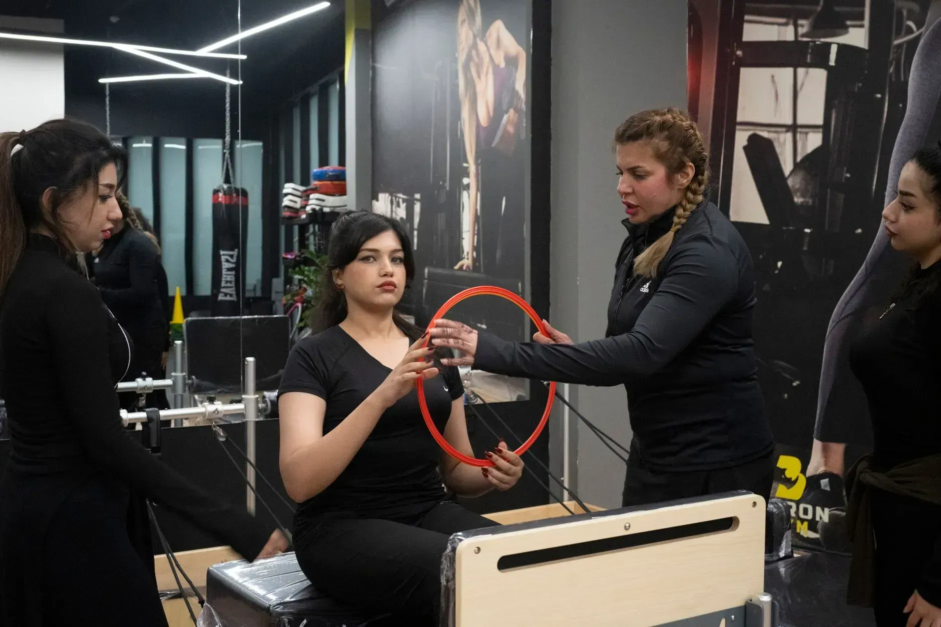 Three women in a gym adjust a seated person’s arm position during a workout session.