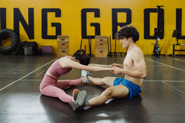 Two athletes stretch on a gym floor, with kettlebells and bold wall lettering in the background.