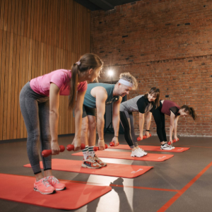 Four people in a gym bending forward with dumbbells on red mats during a fitness class