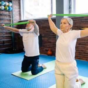 Two people exercising with green resistance bands in a gym studio, one kneeling and one standing on mats